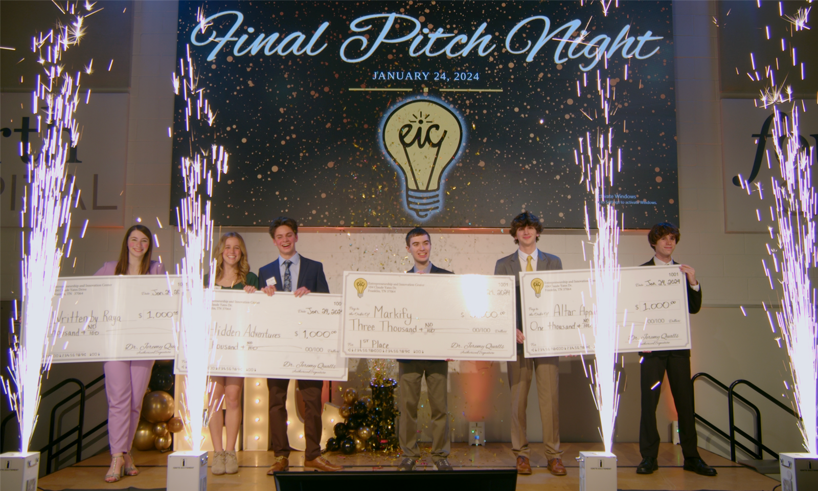 Students stand onstage while holding large paper checks